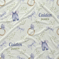 Personalized baby blanket featuring a repeating pattern of lavender baby overalls, wooden ring toys, lavender bows, and green leafy vines on a cream background, with the custom text 'Caiden JAMES' printed in blue and olive green.