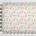 A fitted crib sheet with a repeating pattern of cartoon chickens, chicks, grass, and the name 'Maisie' in orange script, shown inside a white wooden crib.