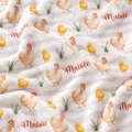 A close-up of a white fabric with a repeating pattern of watercolor-style chickens and chicks in yellow and beige, small green grass tufts, and the word "Maisie" in red script.