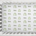 A crib mattress fitted sheet with a repeating pattern of light green cartoon frogs and the custom text 'Kieran ALEXANDER' on a white background, shown inside a white crib.