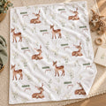 A white fleece blanket with an all-over print of brown deer, small white flowers, and the words 'Dakota james' in green and brown, styled on a woven beige rug with a plush toy and small props nearby.