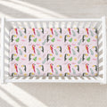 A fitted crib sheet with a colorful toucan and tropical leaf pattern on a light pink background, shown inside a white wooden crib.