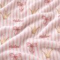 A close-up of a soft plush fabric with a pattern of light pink and white vertical stripes, decorated with watercolor-style pink bows, gold crowns, and the name 'Nadine' in cursive.