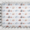 A baby crib mattress fitted sheet with a repeating pattern of colorful toy vehicles (trains, boats, cars, helicopters) and the name 'Oliver' printed in blue script, shown inside a white wooden crib.