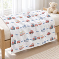 A white baby blanket with a colorful pattern of toy cars, boats, helicopters, and the name 'Oliver' printed in blue, draped over a wooden bassinet with a teddy bear on the corner.