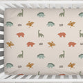 A fitted crib sheet with a dinosaur pattern, shown inside a white wooden crib. The sheet features a repeating pattern of green, orange, and brown dinosaurs on a warm beige background, with the name 'LEO' printed in black between some motifs.
