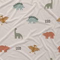 A close-up of a soft fabric surface with an allover dinosaur print on a light warm beige background. The pattern includes sage green long-neck dinosaurs, muted blue-green Stegosaurus-like dinosaurs, and terracotta-brown dinosaurs, with the word 'LEO' printed in black.