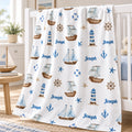 A white baby blanket with a nautical theme, featuring blue and brown illustrations of sailboats, lighthouses, anchors, and stars, along with the name 'Joseph' printed in blue. The blanket is draped over a white wooden crib in a bright, modern nursery setting.