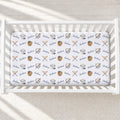 A white crib mattress fitted sheet with a repeating pattern of navy blue 'Jackson' text, baseball caps, baseball gloves, and crossed bats, shown inside a white wooden crib.