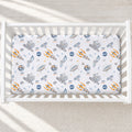 A fitted crib mattress with a white background and a colorful space-themed print featuring rockets, satellites, rockets, and planets. The crib is shown from a top-down view on a light-colored floor with soft shadows.