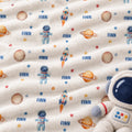 A close-up of a soft fleece blanket with an astronaut and space theme print, featuring the name 'FINN' in blue, rockets, planets, stars, and astronaut motifs on an off-white background. A plush astronaut toy is partially visible in the bottom right corner.