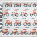A crib sheet featuring a repeating pattern of red-orange watercolor tractors with blue tires and the name 'JOSEPH' printed underneath each tractor, shown inside a white wooden crib.
