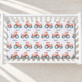 A white crib mattress fitted sheet printed with a repeating pattern of orange-red tractors with blue wheels and small black text, shown inside a white crib on a light neutral floor.