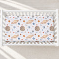 A white crib mattress fitted with a space-themed sheet featuring colorful illustrations of planets, stars, and the name 'Oliver' printed repeatedly, shown in a softly lit nursery setting.
