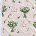 A close-up of a light blush pink fabric with a repeating watercolor print of lily of the valley flower bouquets and the personalized text 'Lillian Grace' in dusty rose and green.
