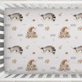 A crib fitted sheet with a repeating woodland print featuring raccoons, hedgehogs, flowers, and the name 'Avery' in a nursery setting.