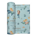 A rolled personalized swaddle blanket featuring a light blue mermaid-themed pattern with illustrations of mermaids, sea turtles, octopus, and starfish, along with the custom text 'ANNA catherine' in gold and dark blue script.