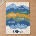 Oliver affirmation blanket laid flat on a wood floor, with seven layered mountain silhouettes, white encouraging phrases, and a large navy name on cream.