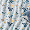 A close-up of a plush blanket or fabric featuring an underwater print with whales, jellyfish, turtles, narwhals, and the word Grace repeated in script. A blue-and-white plush whale toy is partially visible on the right side.