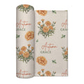 Cream swaddle blanket with golden-orange marigold clusters, green leaves, and the name Autumn Grace in orange and green.