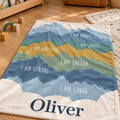 Personalized mountain blanket pairs layered peaks in blues, greens, and warm ochre with your child’s name and seven affirmations: I am kind, I am brave, I am smart, I am grateful, I am enough, I am strong, and I am loved.
