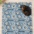 A black and tan dachshund lies curled up on a blue blanket printed with white paw prints and the name Winston repeated across the surface, photographed on a light wood floor.
