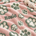 A close-up of a soft pink plush blanket with green paw prints and the name 'Rosie' in dark green lettering. A cream-colored rope toy is partially visible at the top right.