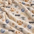 A close-up of a plush cream-colored blanket with a dog-themed print featuring the word 'SPOT', bones, paw prints, bowls, hearts, and blue balls. A knotted rope toy is partially visible in the top-left corner.