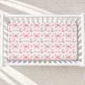 A white crib mattress fitted sheet with a repeating pattern of pink butterflies and the name 'Emery' in script, shown inside a white wooden crib on a light textured rug.