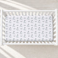 A white crib mattress fitted sheet with a repeating pattern of penguin illustrations and the name 'WESLEY' printed in black, shown inside a white wooden crib.