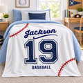 A personalized baseball-themed blanket draped over a bed in a bright, modern bedroom. The blanket is white with navy blue text reading 'Jackson' and 'BASEBALL', the number '19' in navy and white, and red baseball-stitch graphics.