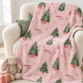 A pink fleece throw blanket with a Christmas tree pattern draped over a cream armchair, styled with a plush bunny toy and a small gift box on a light-colored floor.
