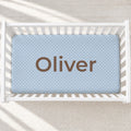 A light blue baby crib mattress cover with white polka dots and the name 'Oliver' printed in brown, shown inside a white wooden crib.