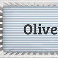 A personalized baby crib sheet with light blue and white horizontal stripes, featuring the name 'Oliver' printed in large black serif letters, shown inside a white wooden crib.