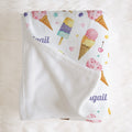 A white baby blanket with a colorful watercolor ice cream and popsicle print, featuring the name 'Abigail' in purple script, laid flat on a white faux fur background.