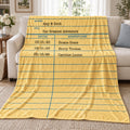 A yellow fleece library card blanket with a lined notebook paper design and printed text fields, draped over a beige sofa in a living room setting.