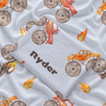 Close-up of a folded Monster Trucks swaddle blanket showing Ryder lettering and watercolor trucks with oversized taupe tires on pale blue.