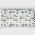 A crib sheet featuring a repeating pattern of vintage toy trains, small houses, trees, and the names 'HENRY' and 'william' on a pale blue background, shown inside a white crib.