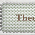 A crib sheet featuring a repeating pattern of small green and white triangles with the partial brown text 'Theo' printed across it, shown inside a white wooden crib.