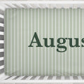A close-up of a crib mattress cover with vertical white stripes on a sage green background, featuring the partial name 'August' in dark green serif lettering.