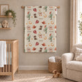 A western-themed nursery wall hanging tapestry with a repeating 'SCOUT' pattern of cacti, cowboy hats, boots, and horseshoes, displayed in a styled nursery room next to a wooden crib and a beige armchair.