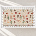 A fitted crib mattress with a western-themed Scout pattern, shown inside a white wooden crib. The pattern features cacti, cowboy hats, boots, horseshoes, and stars in earthy brown, orange, and green tones on a cream background, with the word 'SCOUT' printed repeatedly.
