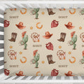 Overhead view of a Wild West crib sheet personalized with Scout, featuring cowboy boots, hats, cacti and bandanas inside a white crib.