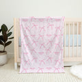 A light pink baby blanket with a repeating pattern of pink bows and the custom text 'Charlotte MAE' is draped over a light wood baby crib in a nursery setting.