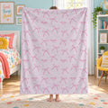 A child standing in a bedroom holding up a large light pink blanket or throw featuring a repeating pattern of pink bows and small pink stars, with the words 'Charlotte MAE' printed inside some bows.