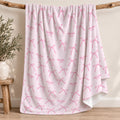 A light pink fleece blanket with a repeating pattern of pink bows and the names 'Charlotte MAE' in script, draped over a rustic wooden branch stand in a styled room setting.