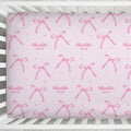 Personalized blush pink crib sheet with watercolor bows, tiny sparkles, and the repeated name Charlotte Mae, shown inside a white crib.