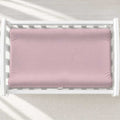 A dusty pink changing pad cover inside a white wooden baby changing table, shown from a top-down view on a light textured floor with soft diagonal shadows.