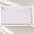 A top-down view of a white wooden crib with a fitted sheet featuring a light pink and white chevron pattern, set against a neutral cream background with soft diagonal shadows.