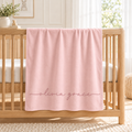 A light pink baby blanket draped over a wooden crib, featuring the name 'Olivia grace' embroidered in a matching dusty rose script at the bottom.